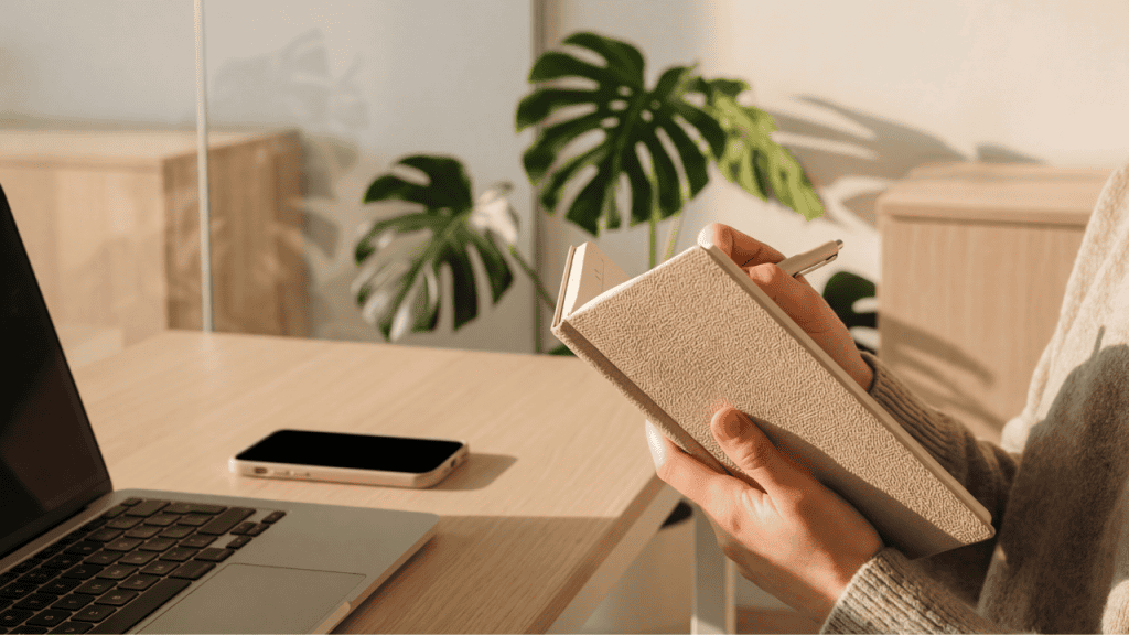 Primer plano de manos escribiendo en una libreta sobre un escritorio de madera clara, con una laptop y un smartphone a un lado. Al fondo, una planta monstera bajo una luz natural cálida y estética minimalista.