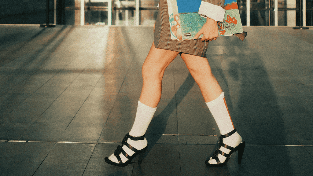 Woman walking on pavement with white socks and black heeled sandals, wearing a blazer.