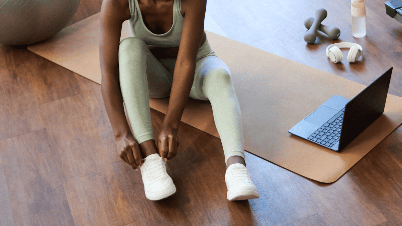 Mujer en ropa deportiva sentada sobre un mat de ejercicio en casa, atándose las zapatillas deportivas junto a una computadora portátil en una mañana iluminada.